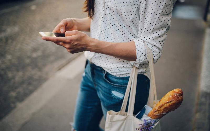 Close to the center of everything woman walks down the street and texts with a bag of groceries on her arm