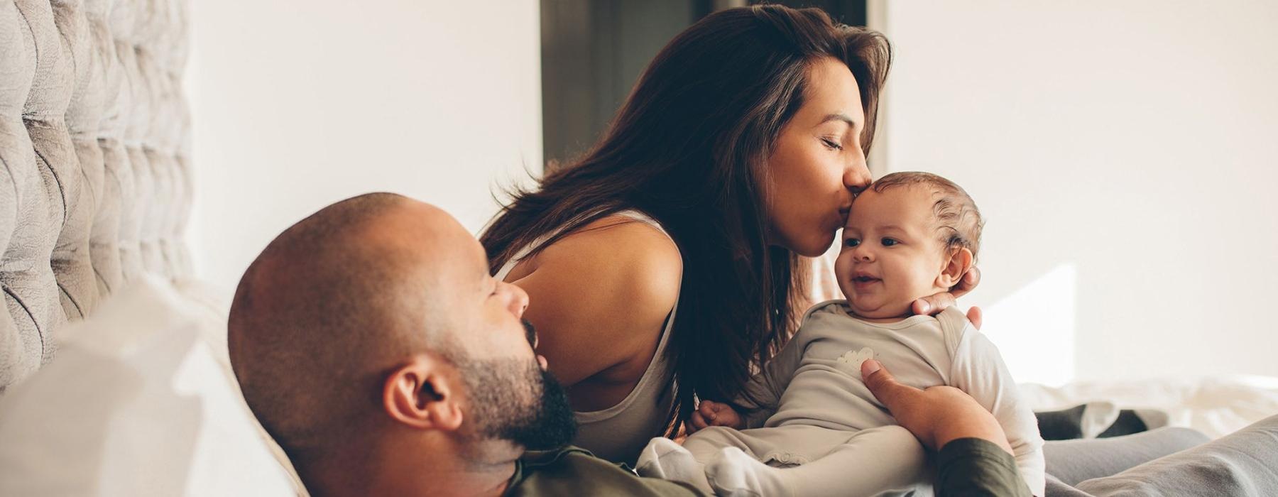mother kisses her infant while father lies in bed and holds him